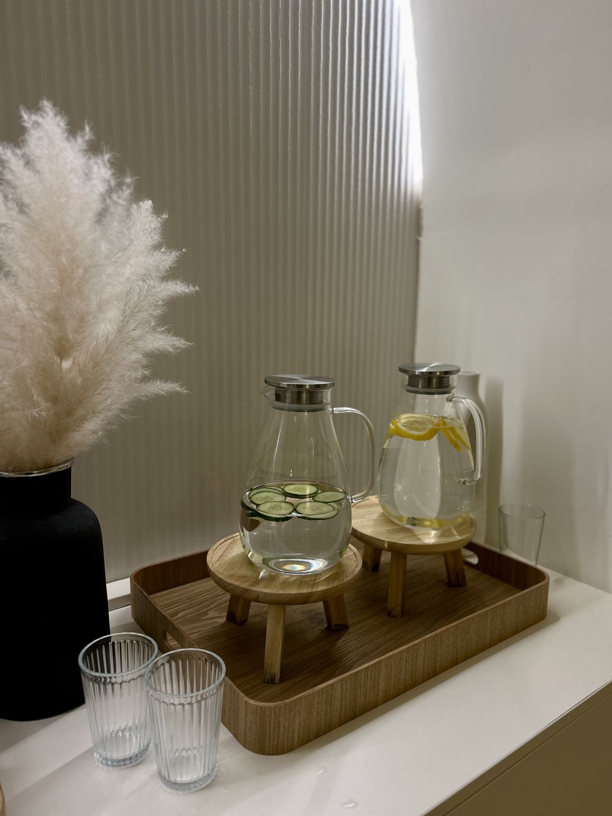 A wooden tray with glass jugs of cucumber and lemon water, set beside pampas grass in a quiet reception corner.